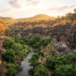 Panoramic view of ancient rock-cut cave temples carved into cliff faces along a winding river with trees and visitors exploring the site.